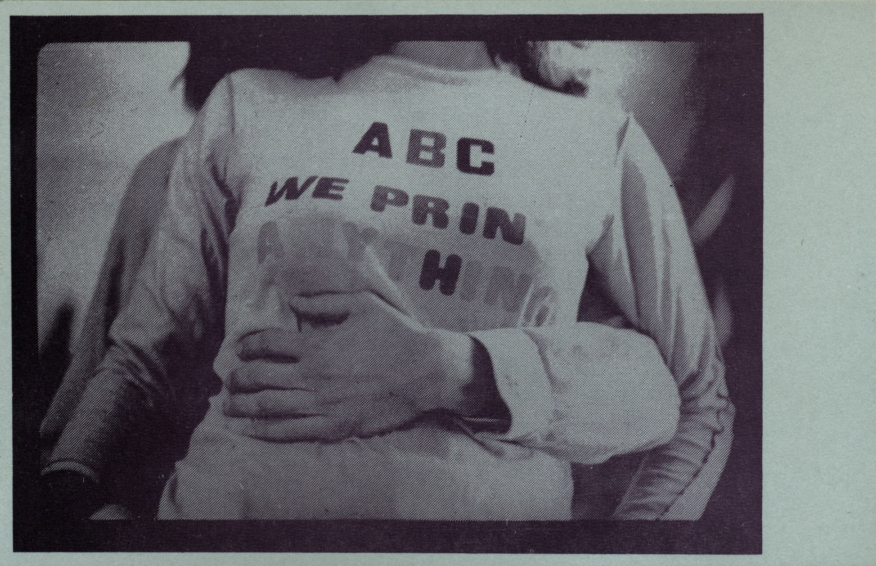 black and white image zoomed in on someone's shirt which says "ABC We Print Anything;" Another person's arm is wrapped around them
