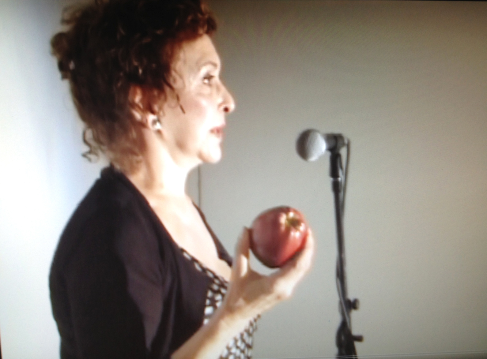 a woman stands in profile view, an apple in her hand, speaking into a microphone