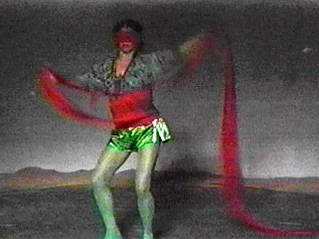 image of a figure in green tights and metalic green shorts; a long, red strip of fabric is wrapped around their waist and eyes, and is held in both of the figure's hands.