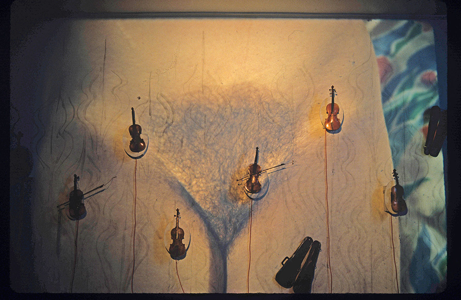 six violins are installed over a projected image of a woman's nude crotch standing in front of a blue, red, and white abstract background