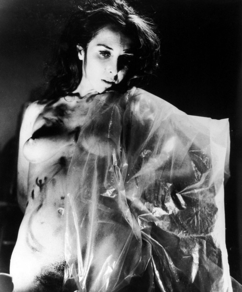 Black and white photo of artist standing naked partially covered in transparent plastic sheeting.