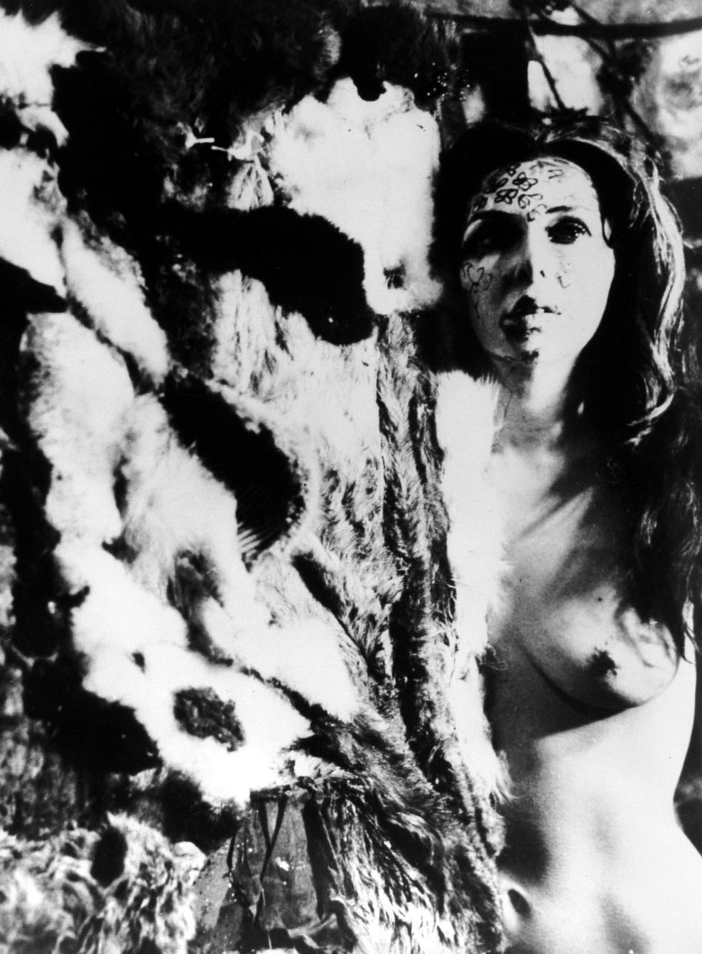 Black and white photo of the artist standing naked from the waist up, the left half of her body is obscured by materials and there is paint on her face.