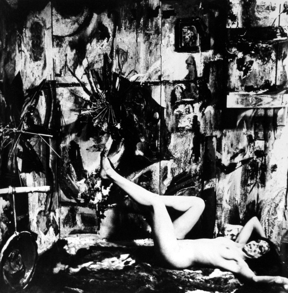 Black and white photo of artist reclining naked in front of paintings.