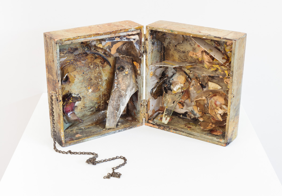 open light wooden box filled with shattered glass and paint splatters; a metal chain curls around from the left side to in front of the box