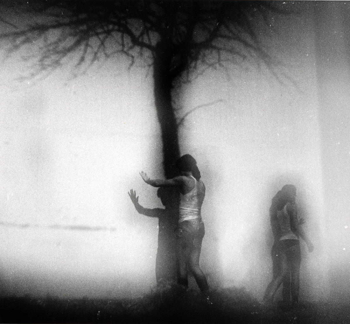 black and white image of two full body figures, one faces a black projection of a tree