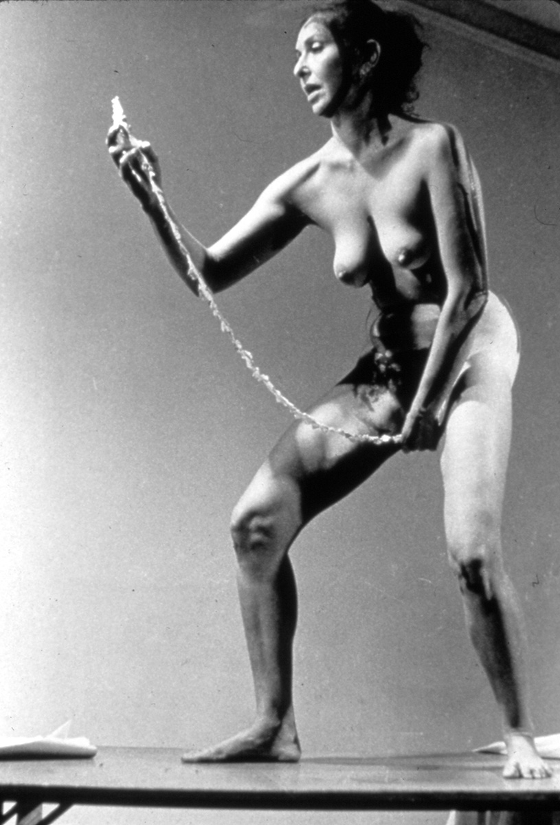 b/w photo of Schneemann mid performance; she is nude and slightly hunched over; she is removing a thin paper scroll from her vagina and reading it