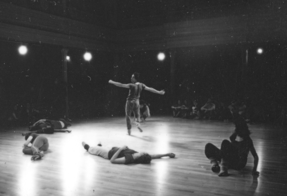 black and white photo of several figures lying on a wooden floor; in the center a figure stands with their arms outstretched