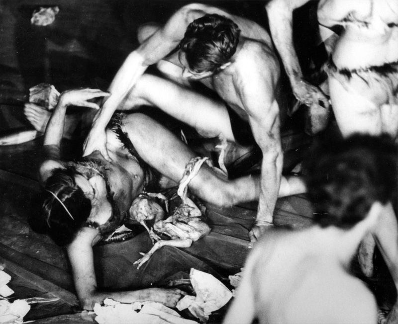 Black and white photo of perfromers on top of each other on the ground, covered in blood and paint. At the center of the photo are two chicken carcasses.
