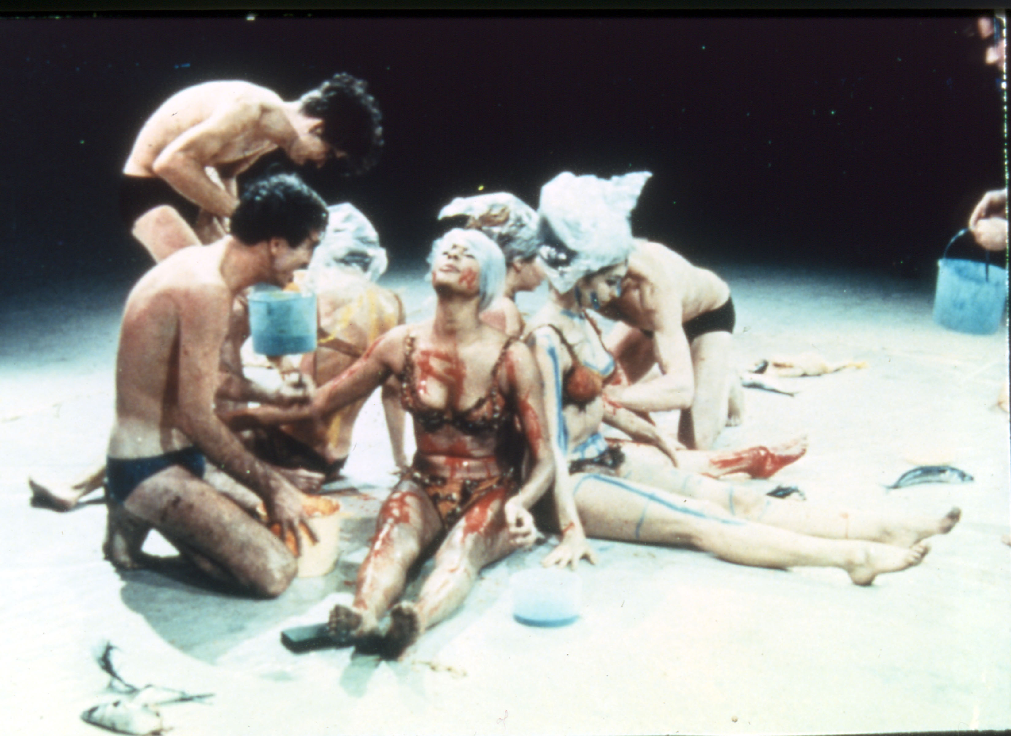 Four female performers sit in their underwear covered in paint and blood and three male performers are crouched facing them.
