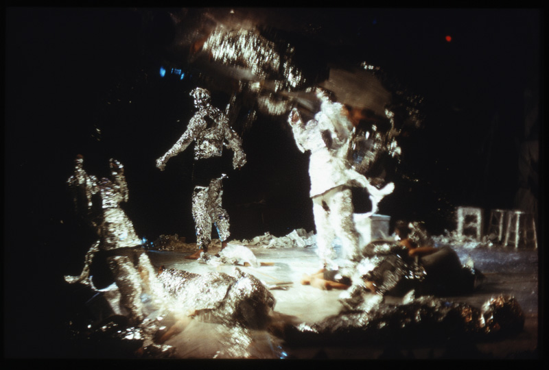 five figures in metalic, tin-foil like outfits in motion under bright white light