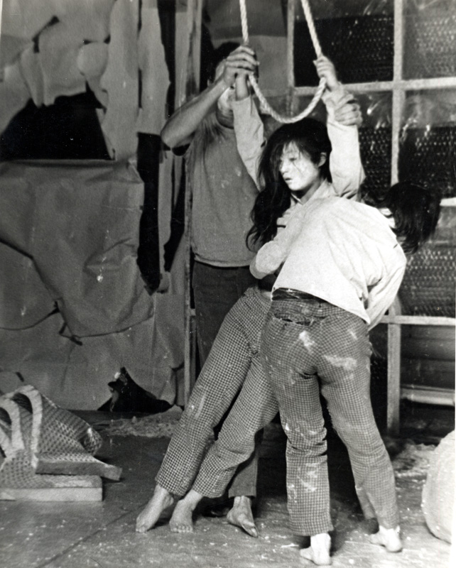 black and white image of three figures wearing plaid pants and white masks, the center figure is hanging from a rope