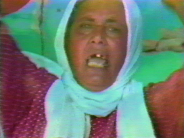 close up of a face with arms rasied and mouth open; the subject is wearing a red shirt with a white pattern and a white scarf wrapped around their head
