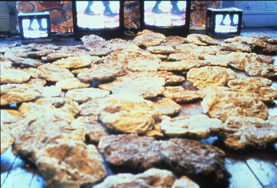 detail of installation of Video Rocks showing hand-cast rocks (cement, glass, ashes, wood) and video monitors on the wooden floor; on the monitors plays two-channel video of feet walking on rocks