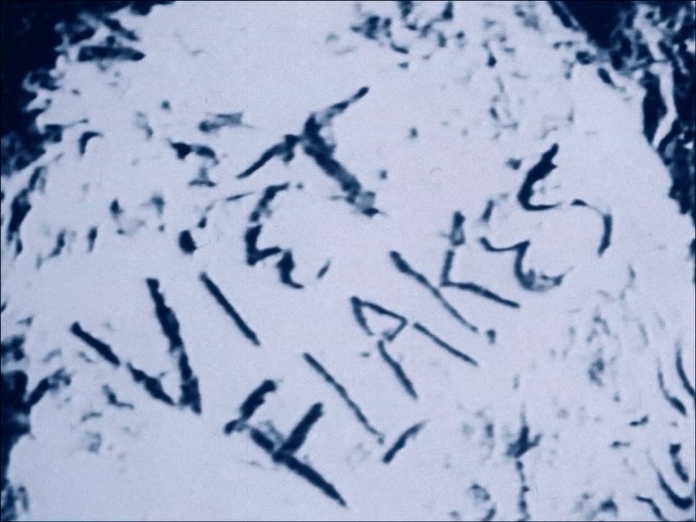 still of several figures crying: one is lying down, another looks directly at the camera, behind them there is a small boy; overlaying the image is a pink filter which abstracts the shot