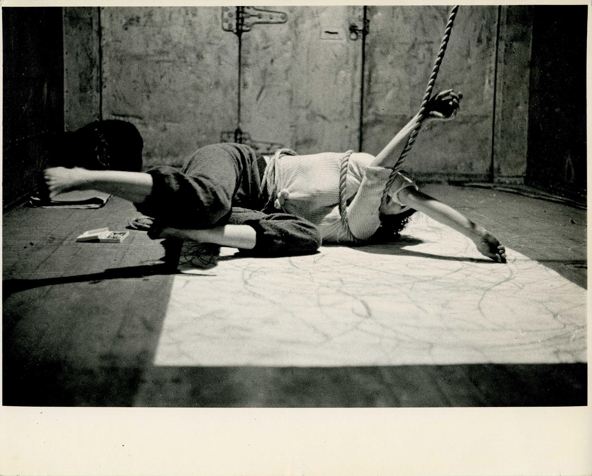 b/w image of the artist laying on the floor, a rope wrapped around her upper body; she is drawing on a large white sheet of paper on the ground