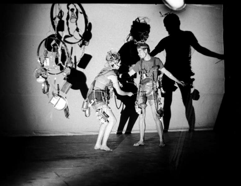 b/w image of man and women on stage with a bright white light casting their shadows behind them; an abstract sculpture of a metal circle with mixed media items hanging from the circle is to the left of the figures